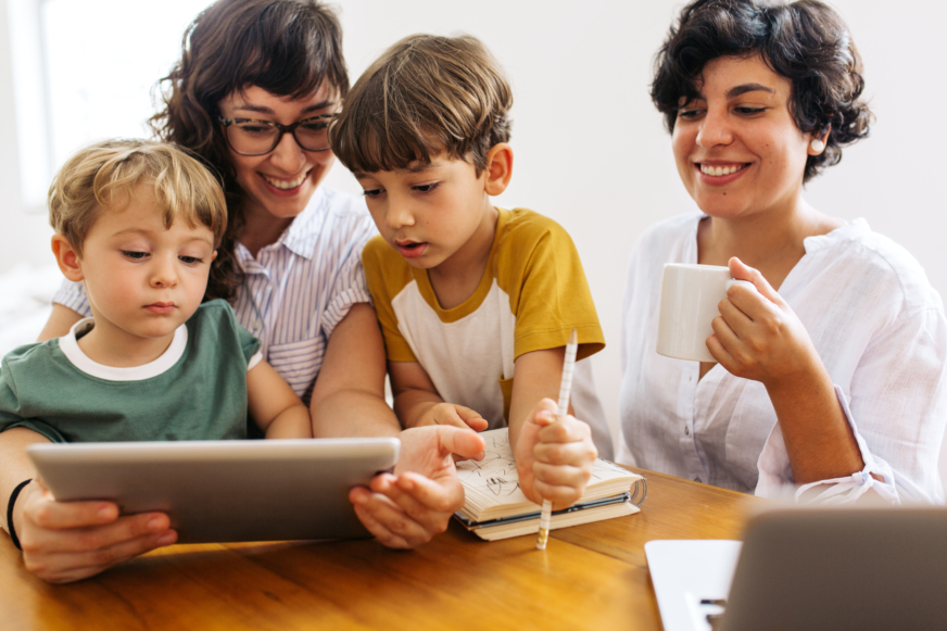 A lesbian couple smiles at an iPad next to their two young children while sitting at a wooden dining table.