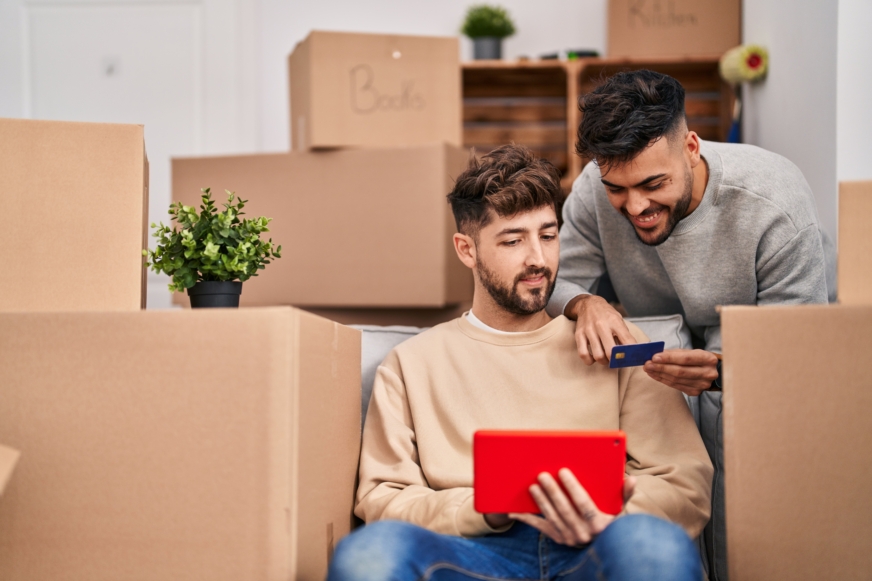 A young gay couple stands smiling around cardboard boxes to be unpacked.  They read the credit card number while working on a tablet screen. 