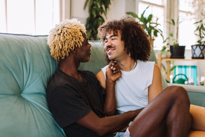 A gay black couple sits on a sofa in their living room and smiles lovingly, looking into each other's eyes.