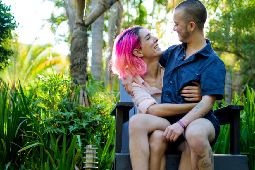 A non-binary couple sits on a patio chair in a beautiful grassy backyard.  They look at each other lovingly.